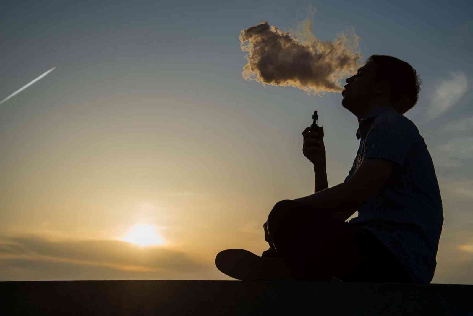 vaping young man with, produces vapor on sunset sky background at the sea coast promenade, place for text.